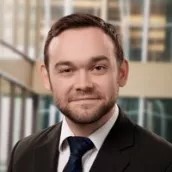 Male with brown hair and beard, wearing black suit and white blouse with blue tie