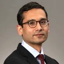 Male wearing eyeglasses, light blue blouse, black blazer and red tie