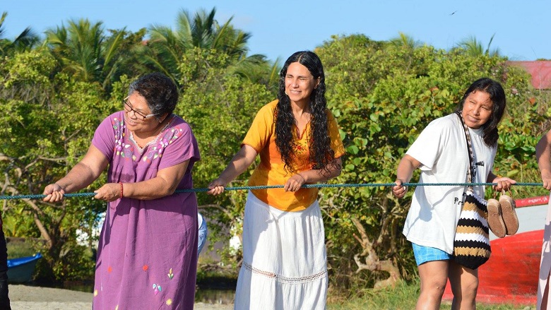 Three women pull on a rope on a beach. The one on the left wearing a pink dress with a floral pattern is Myrna Cunningham.