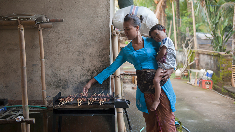 Woman holding a young child tending a grill with cooking meat