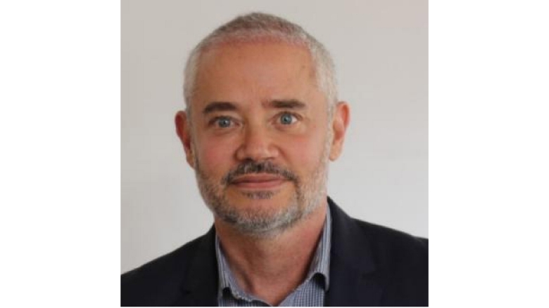 Male with white hair and grey beard, wearing black blazer and grey blouse