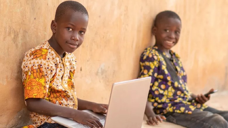 Two Mozambican children each hold a laptop and a cellphone while smiling at the camera