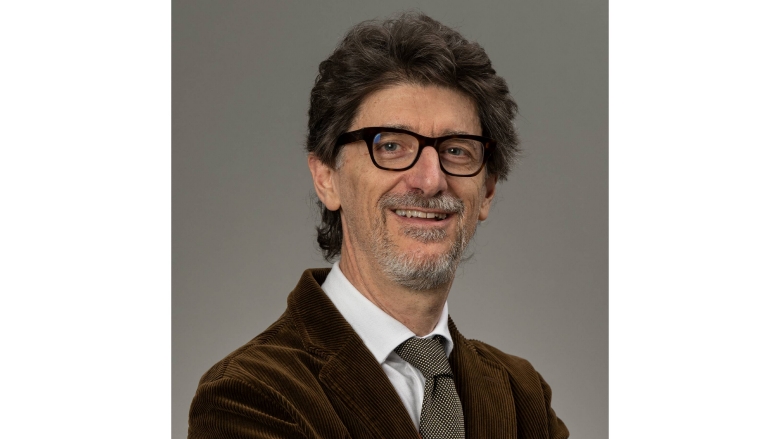 Male with dark brown hair, wearing eye glass, brown blazer, white blouse and pattern tie