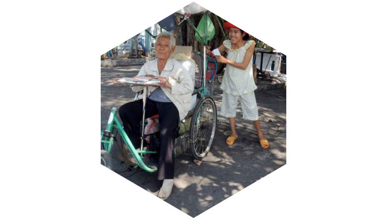 A senior Vietnamese woman in a wheelchair smiles with her grandchild standing behind her